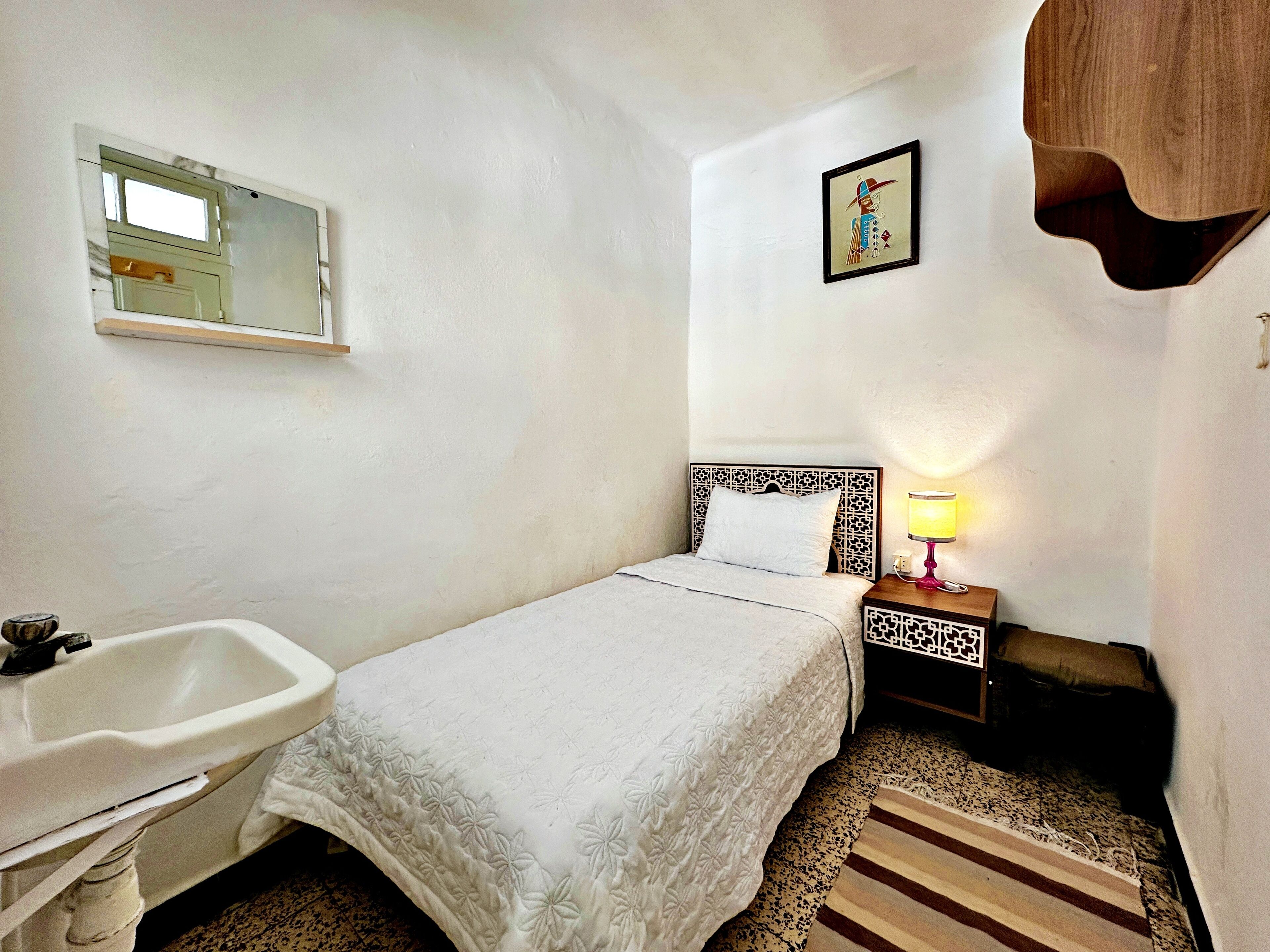 Ein kleines, sauberes Zimmer mit einem Einzelbett, einem Nachttisch mit Lampe, einem kleinen Ablagetisch, einem Spiegel und einem Waschbecken.