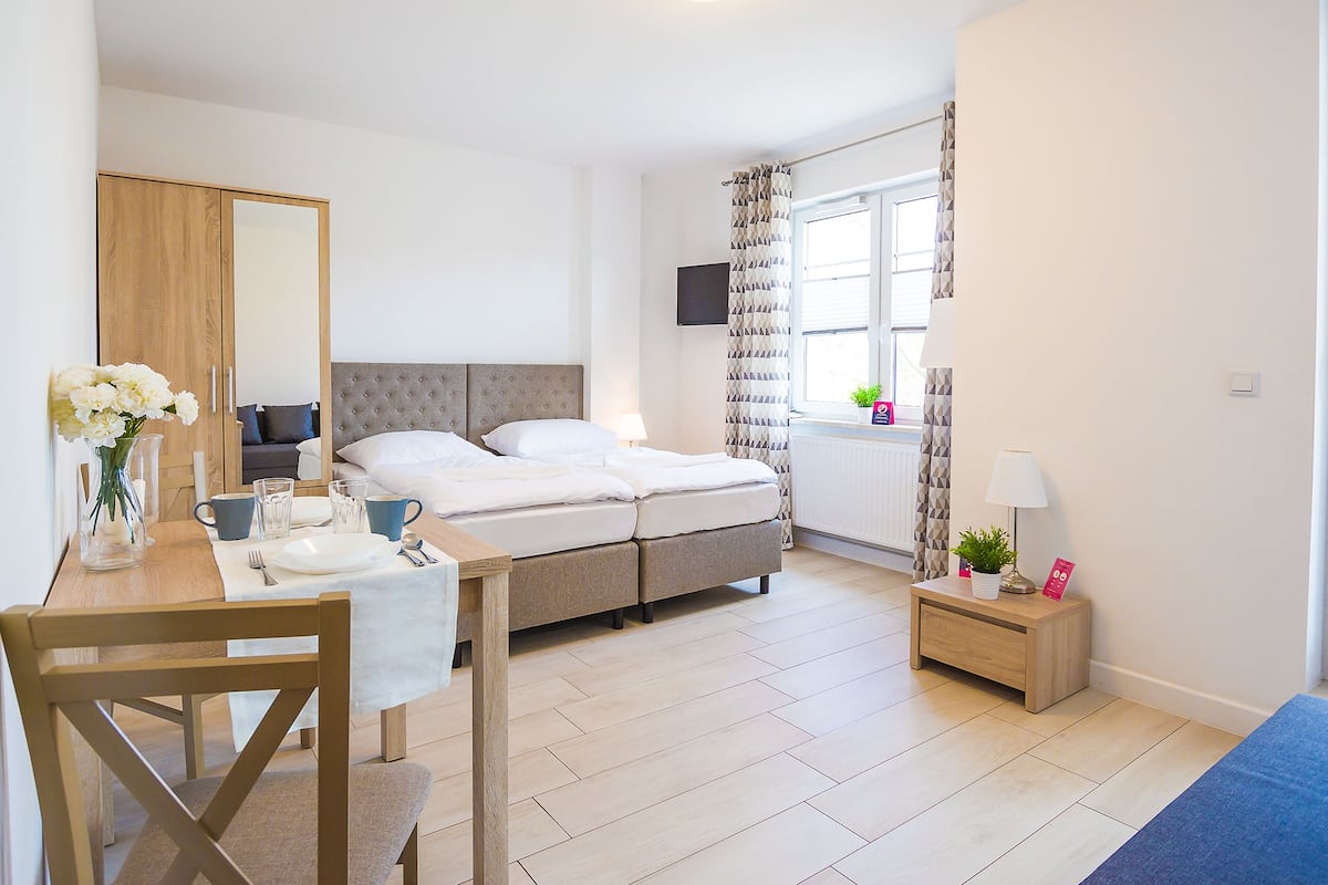 A modern hotel room with a bed, a desk with a chair, a small table with a vase of flowers, and a window with curtains.