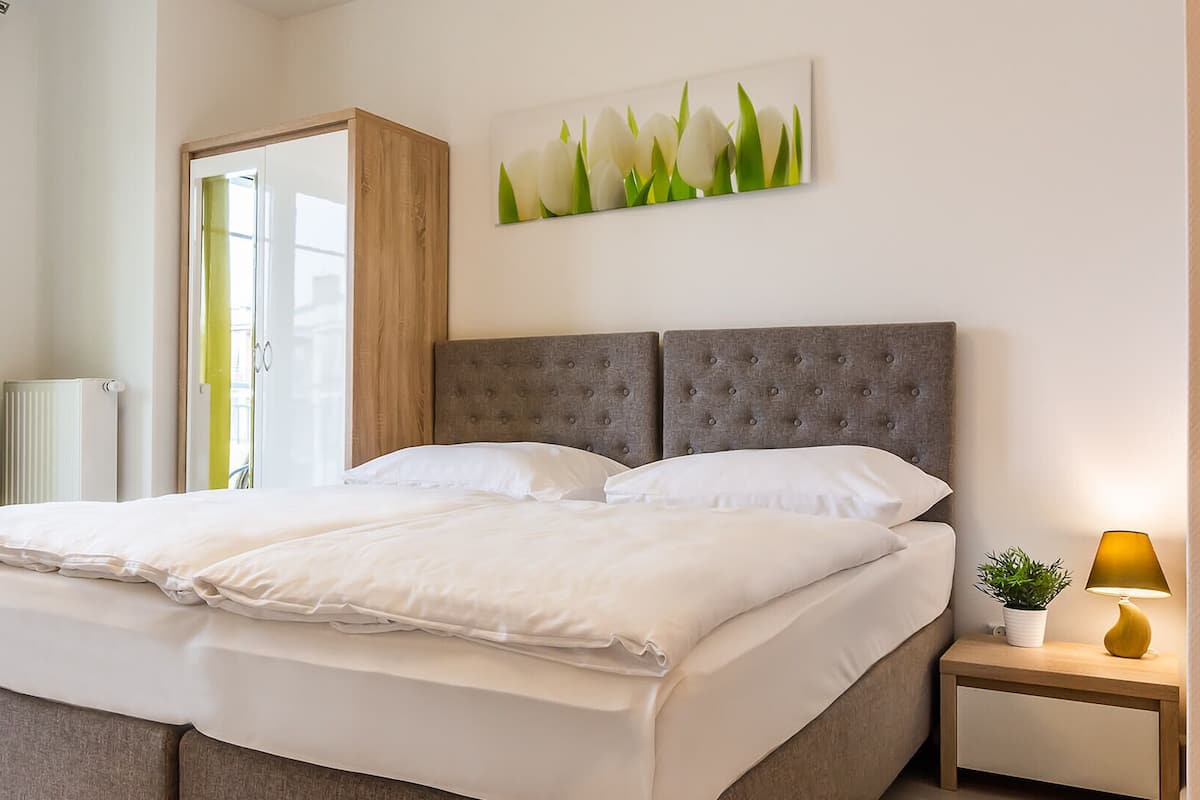 A double bed with a grey upholstered headboard, white bedding, and a bedside table with a lamp and a plant.