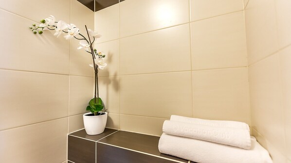 A bathroom with beige tiles, a white orchid in a white pot, and a stack of white towels. - VacationClub - Aquamarina Apartments (Swinoujscie)