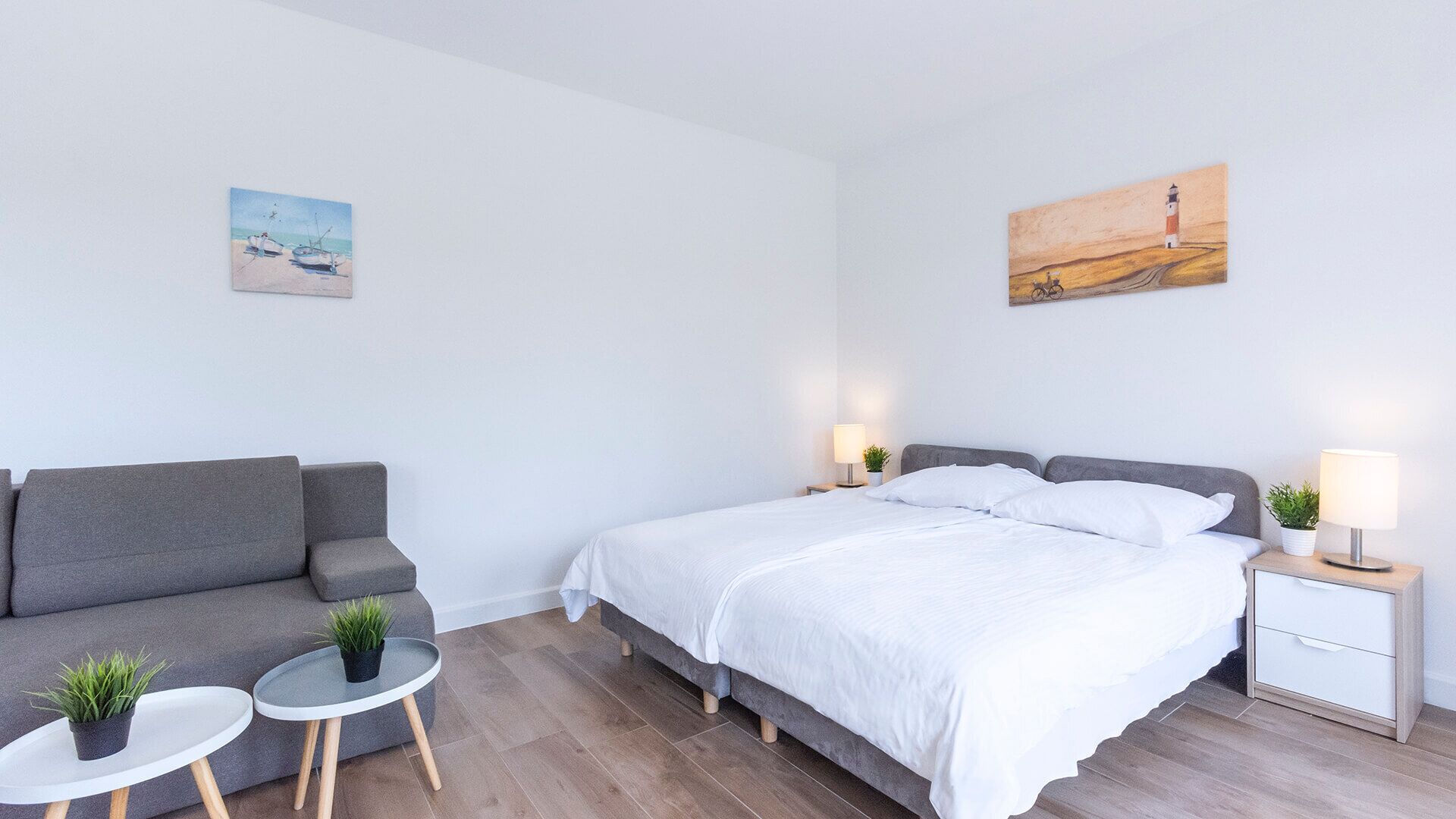 A modern bedroom with a bed, bedside tables with lamps, a grey sofa, and a small table with a plant.