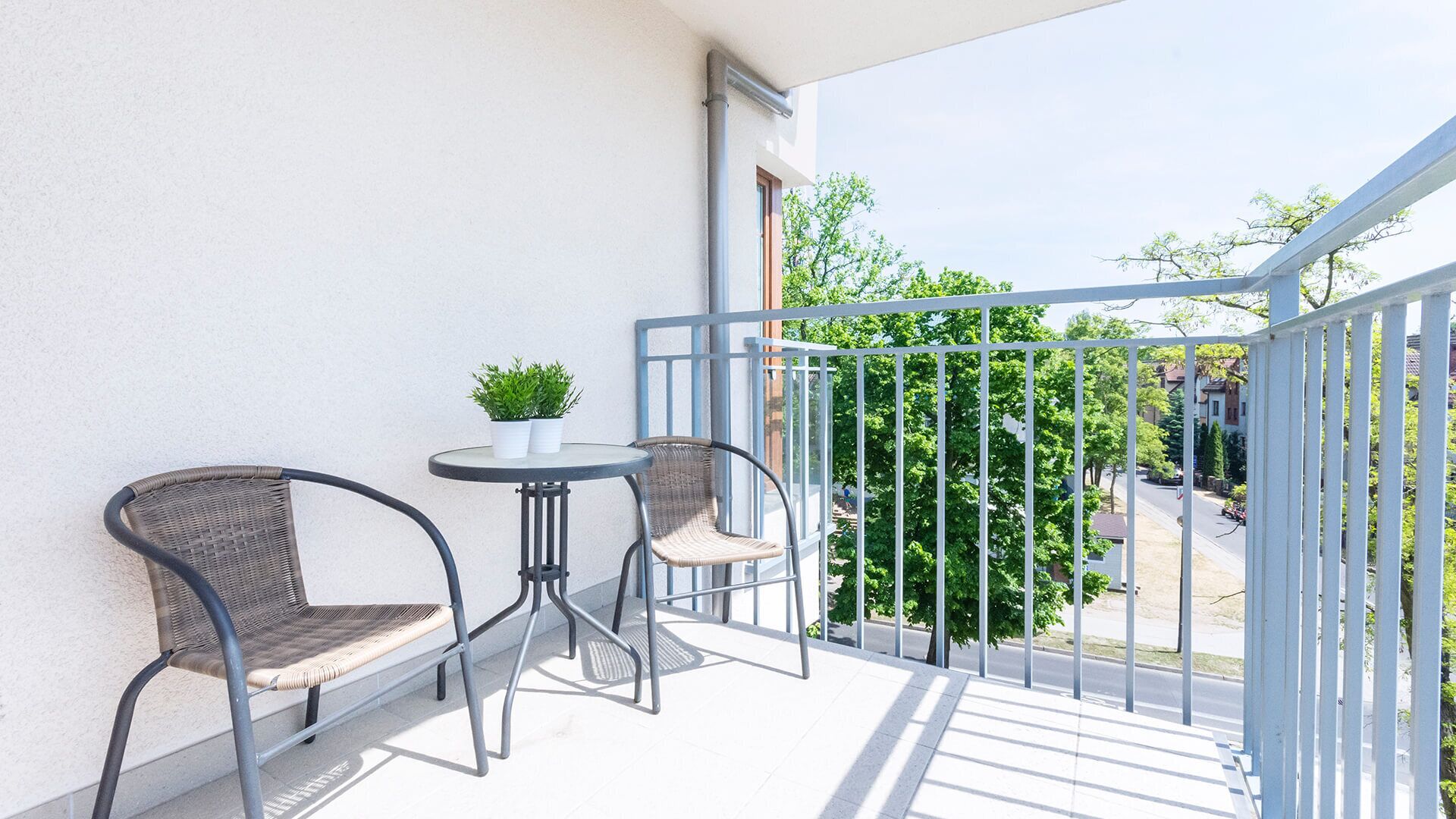 Varanda com duas cadeiras de vime, uma mesa pequena, um vaso de planta e vista para uma rua residencial.