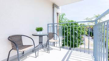 Varanda com duas cadeiras de vime, uma mesa pequena, um vaso de planta e vista para uma rua residencial.