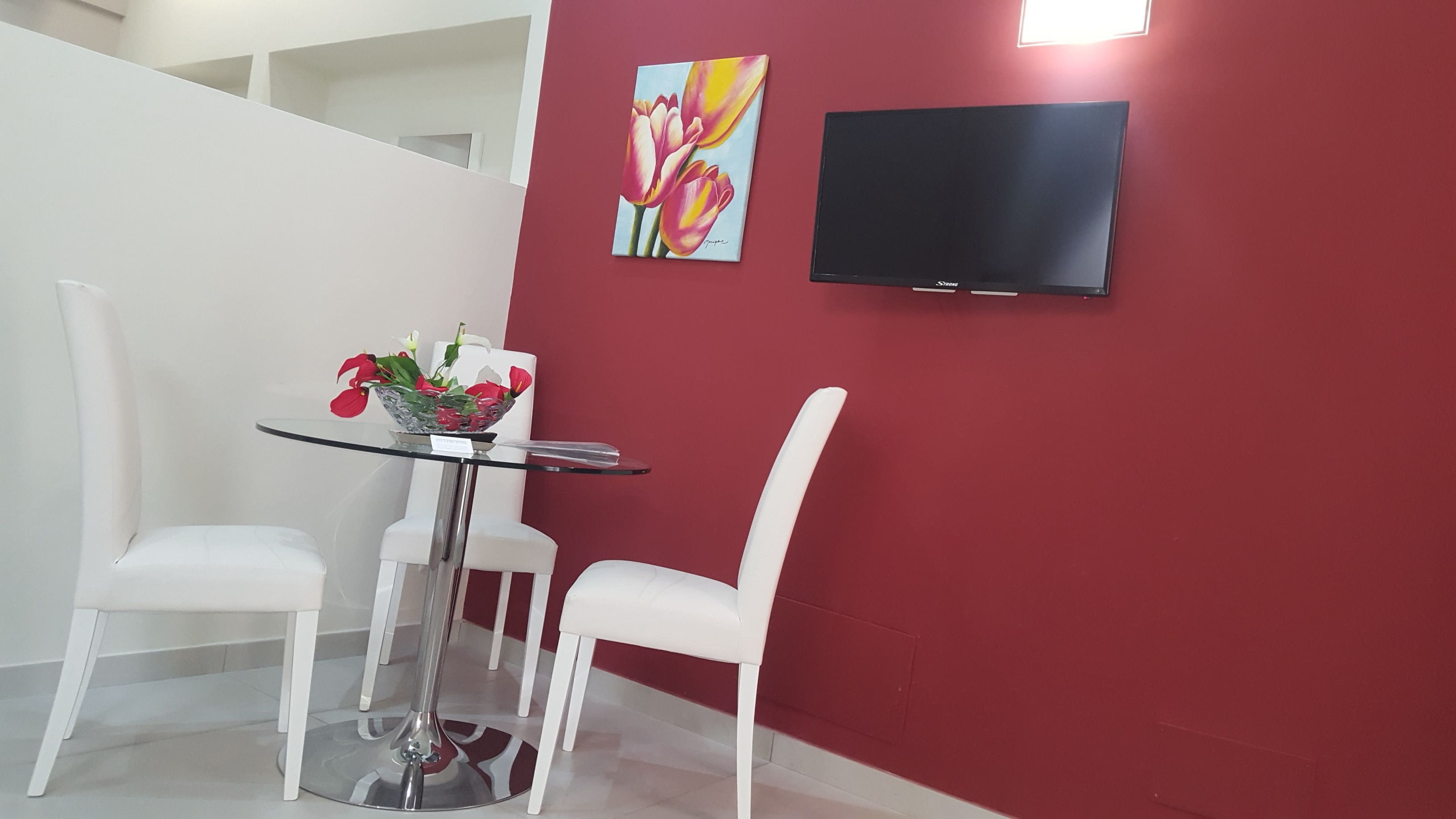 A modern room with a red accent wall, a flat-screen TV, a glass table with chairs, and a floral painting.