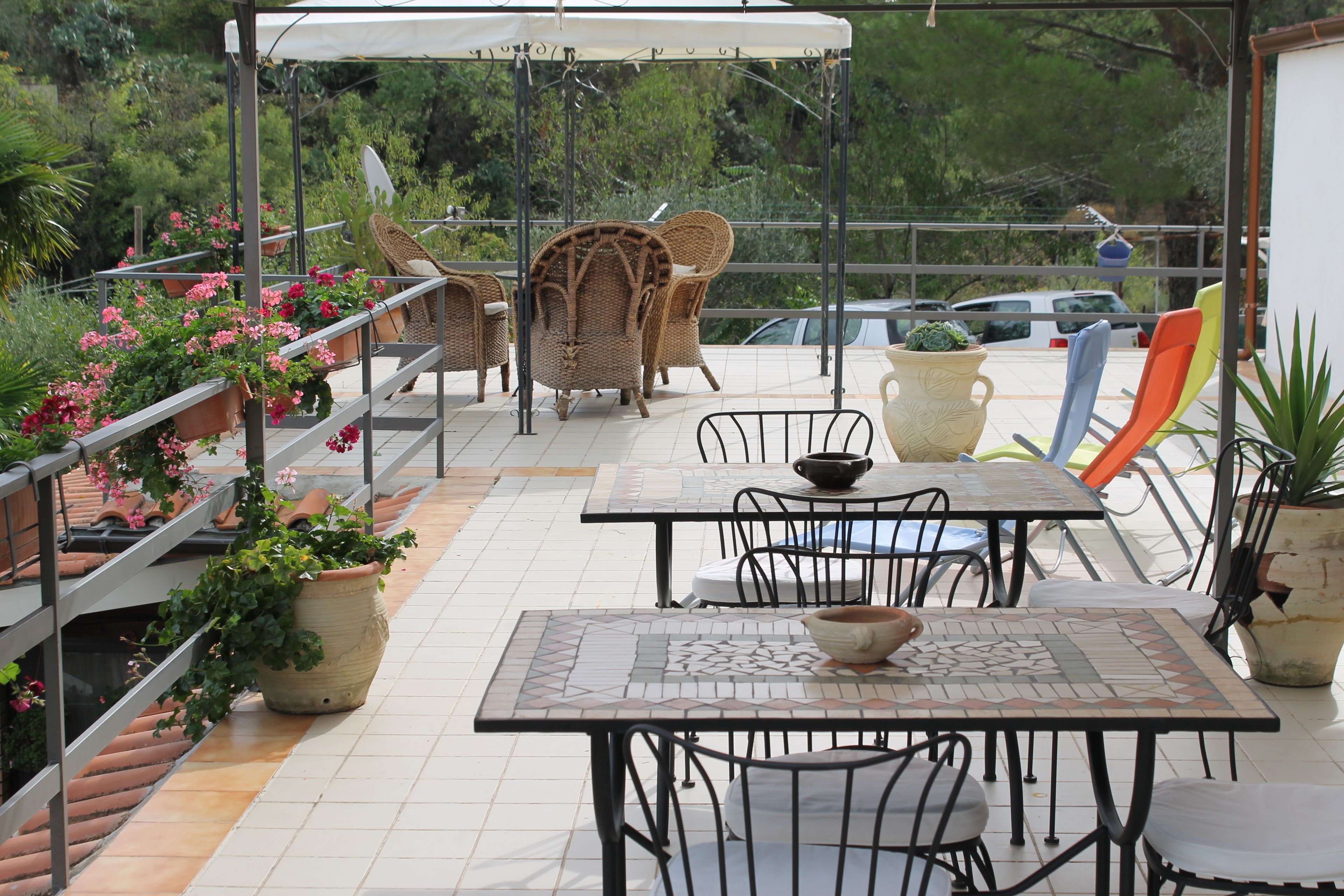 An outdoor patio with tiled flooring, metal tables and chairs, potted plants, and a covered seating area.