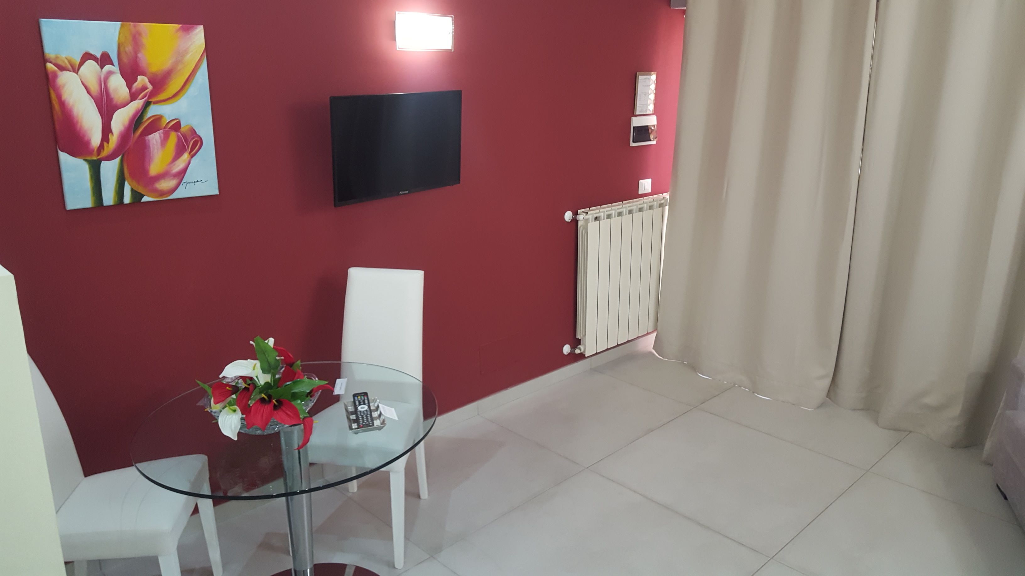 A room with a red wall, a flat-screen TV mounted above a white chair, a glass table with a vase of flowers, and a radiator.