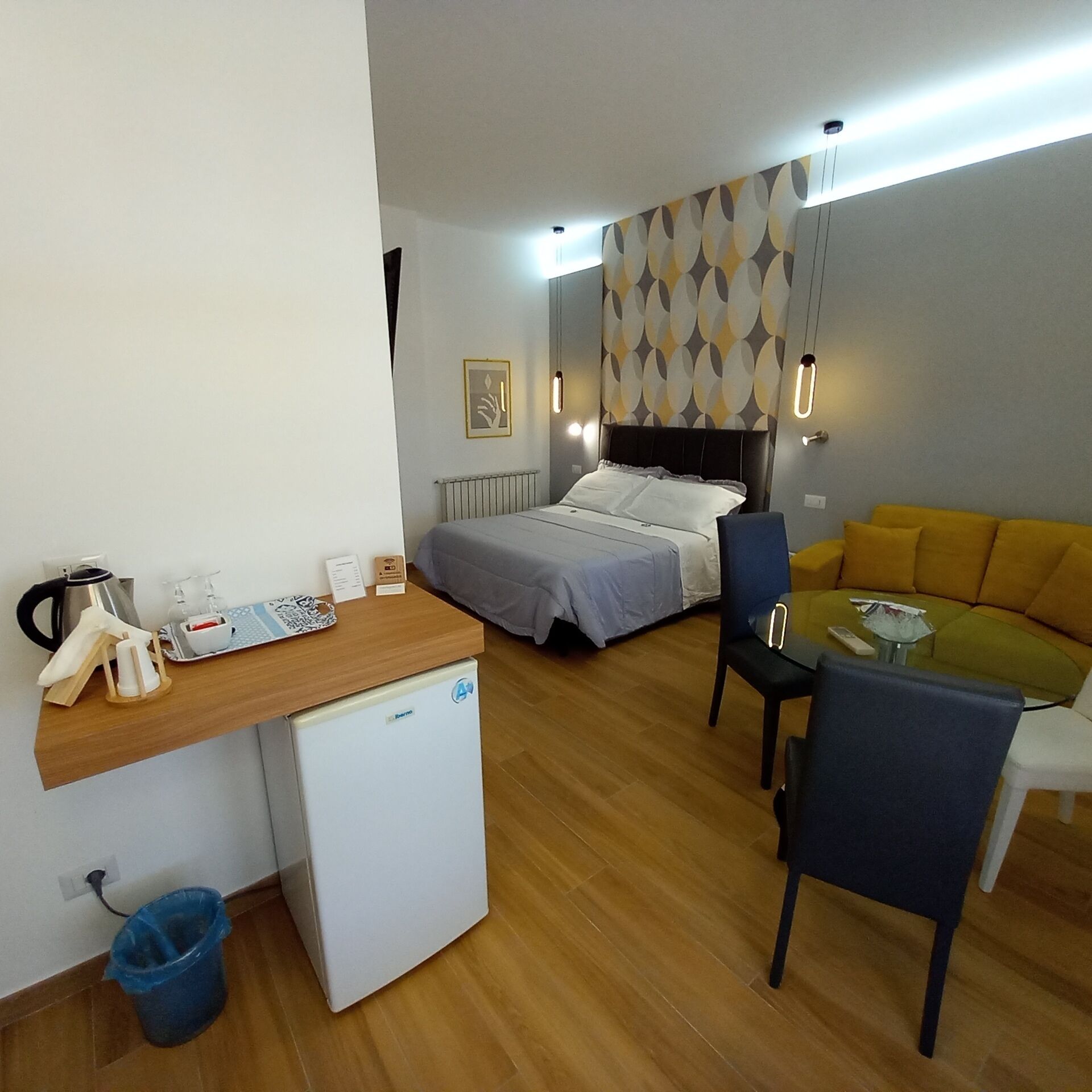 A compact hotel room with a bed, a desk, a chair, a small refrigerator, and a sofa.