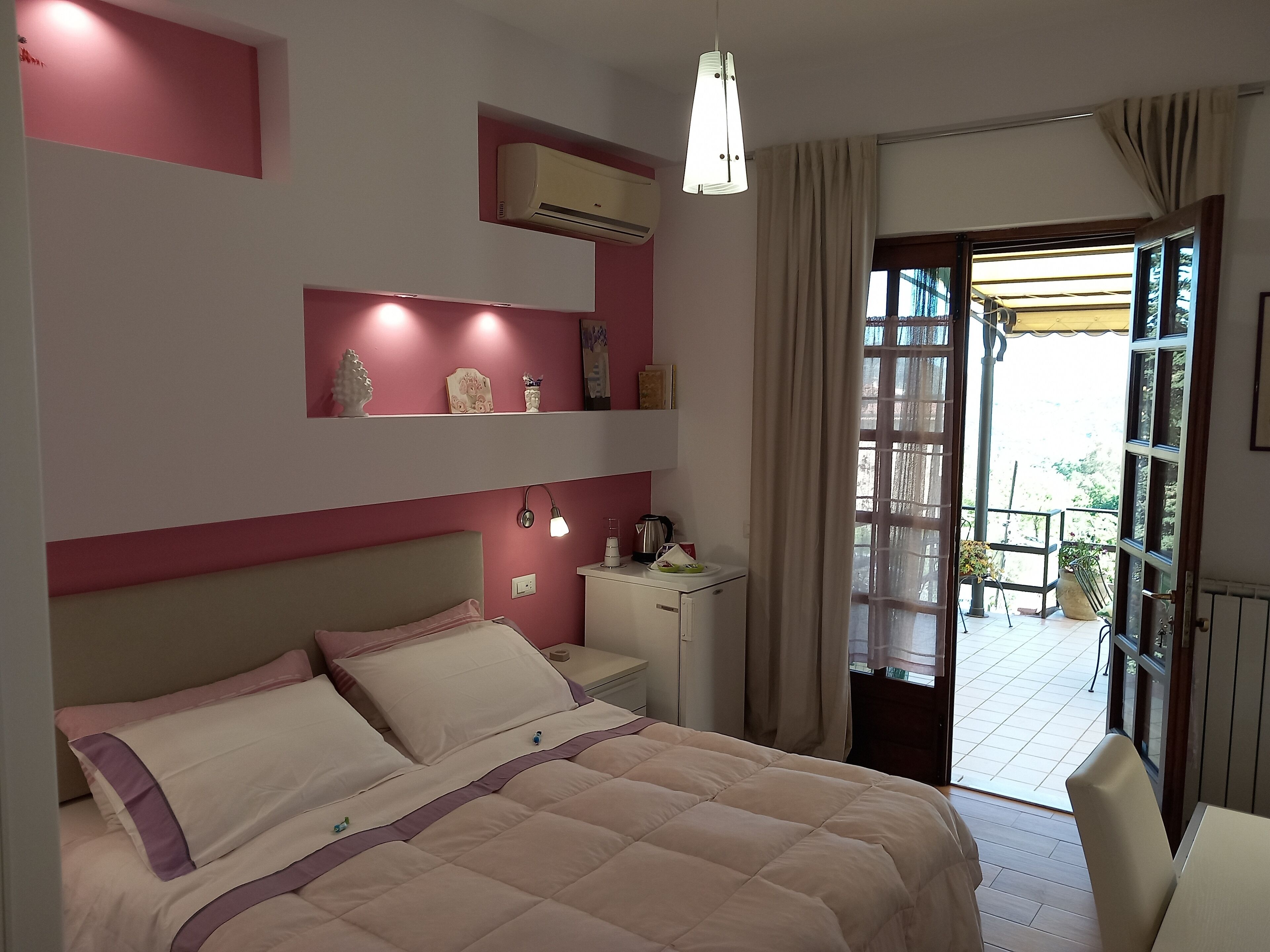 A bedroom with a bed, bedside table, lamp, air conditioning unit, and a view of the outside balcony.