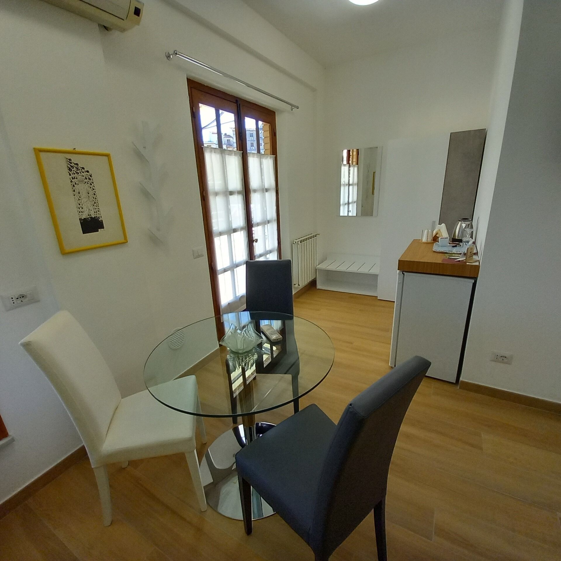 A modern, well-lit room with a glass dining table, white chairs, a small kitchenette, and a large window with blinds.