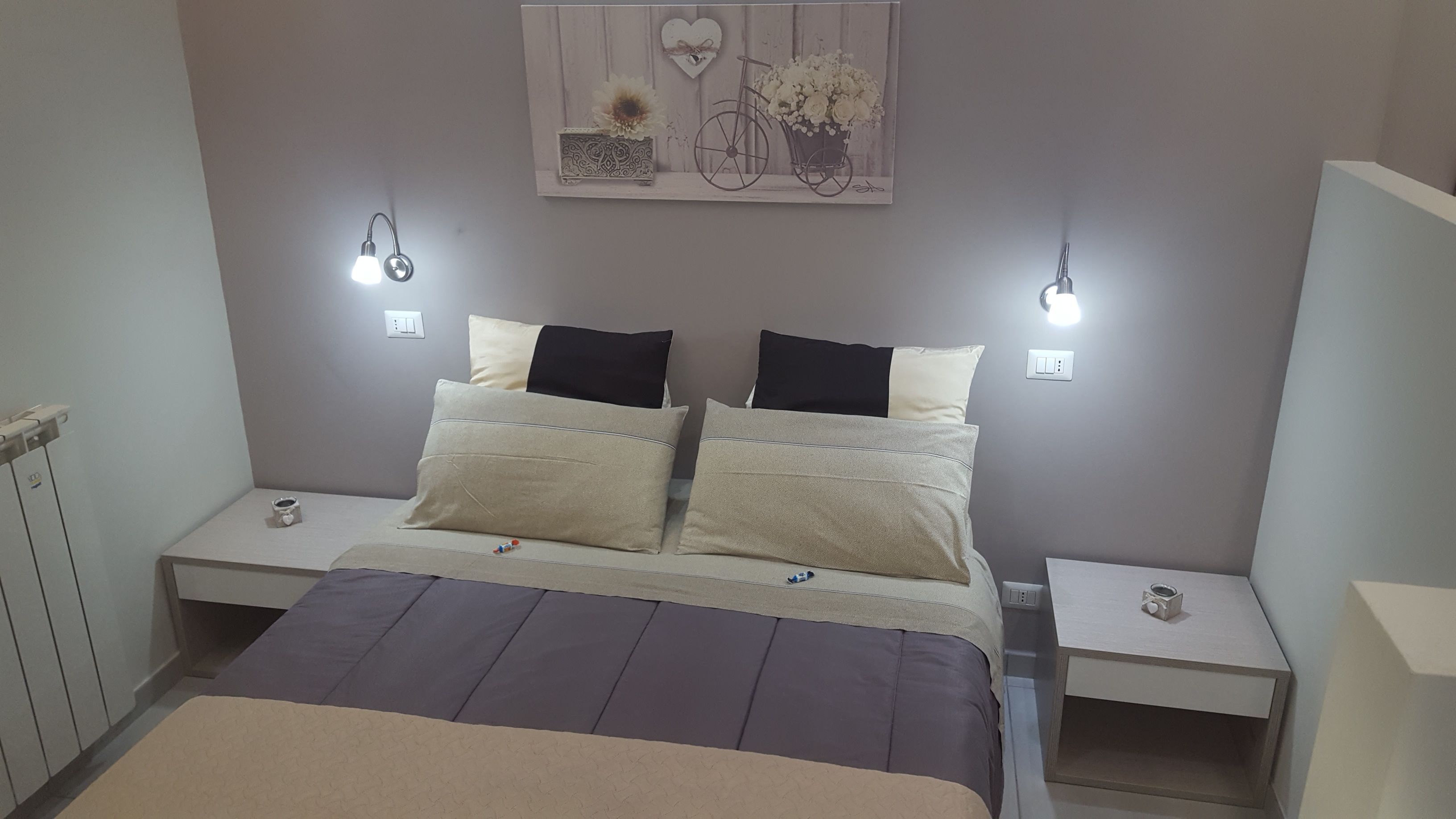 Quarto bem arrumado com cama, duas mesas de cabeceira, um criado-mudo, luminária na parede e um objeto decorativo sobre a cama.