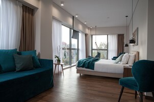 Moderni hotellihuone, jossa on suuri sänky, sohva ja nojatuoli. Huoneessa on useita ikkunoita, joista tulee runsaasti luonnonvaloa ja joista avautuu näkymä kaupunkiin.