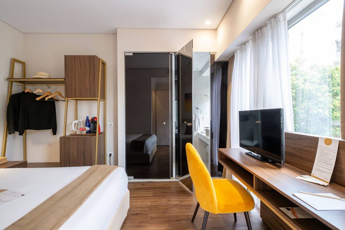 A hotel room with a bed, a desk with a TV, a yellow chair, a wardrobe, and a view of the outdoors.