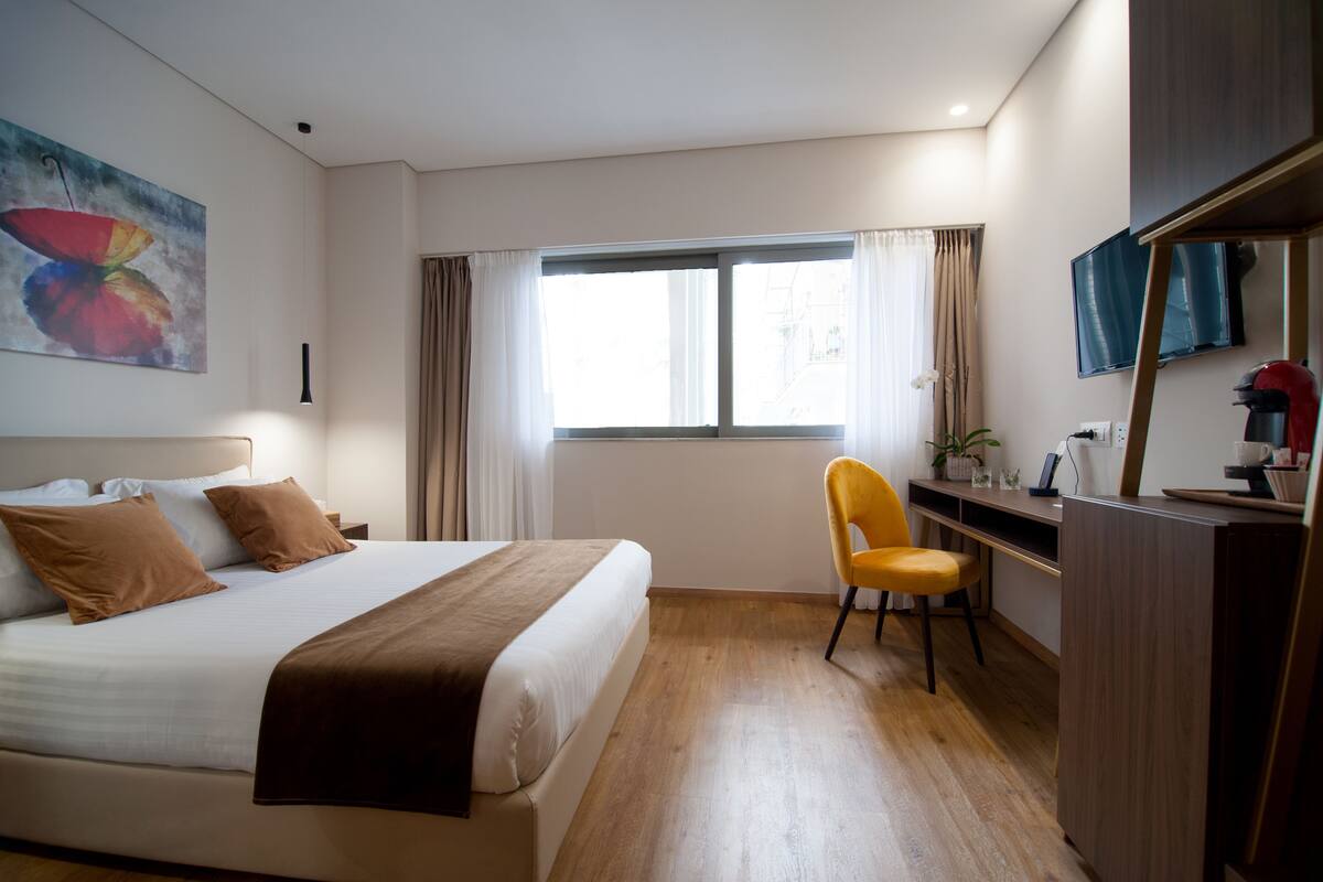 A hotel room with a bed, a desk, a chair, a window, and a painting on the wall.