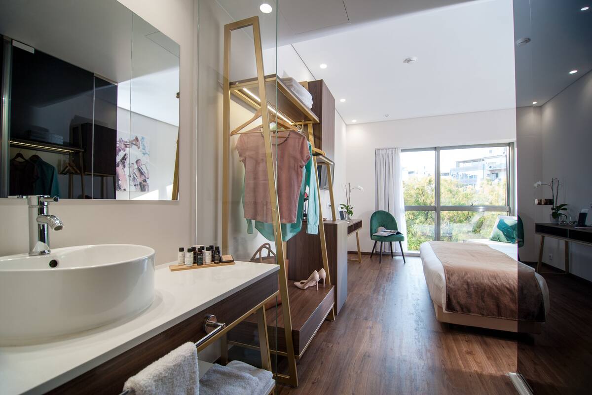 A modern hotel room with a large bed, a bedside table, a desk, a chair, a large window, and a bathroom with a sink and mirror.