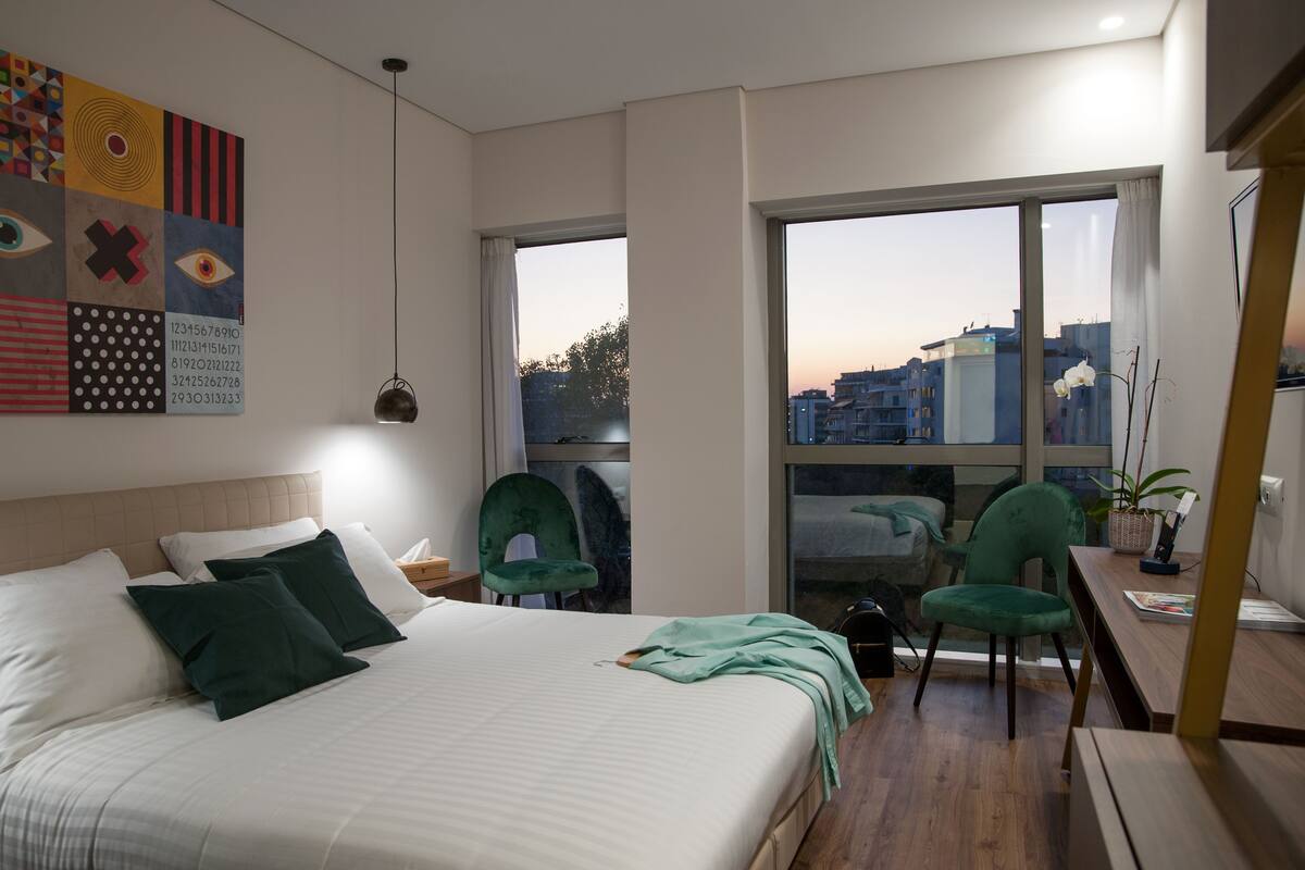 A modern hotel room with a large bed, two chairs, a desk, and a view of the city through large windows.