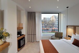 Una camera d'albergo con un letto, una sedia, una scrivania con una lampada, vista su edifici e una finestra con tende.