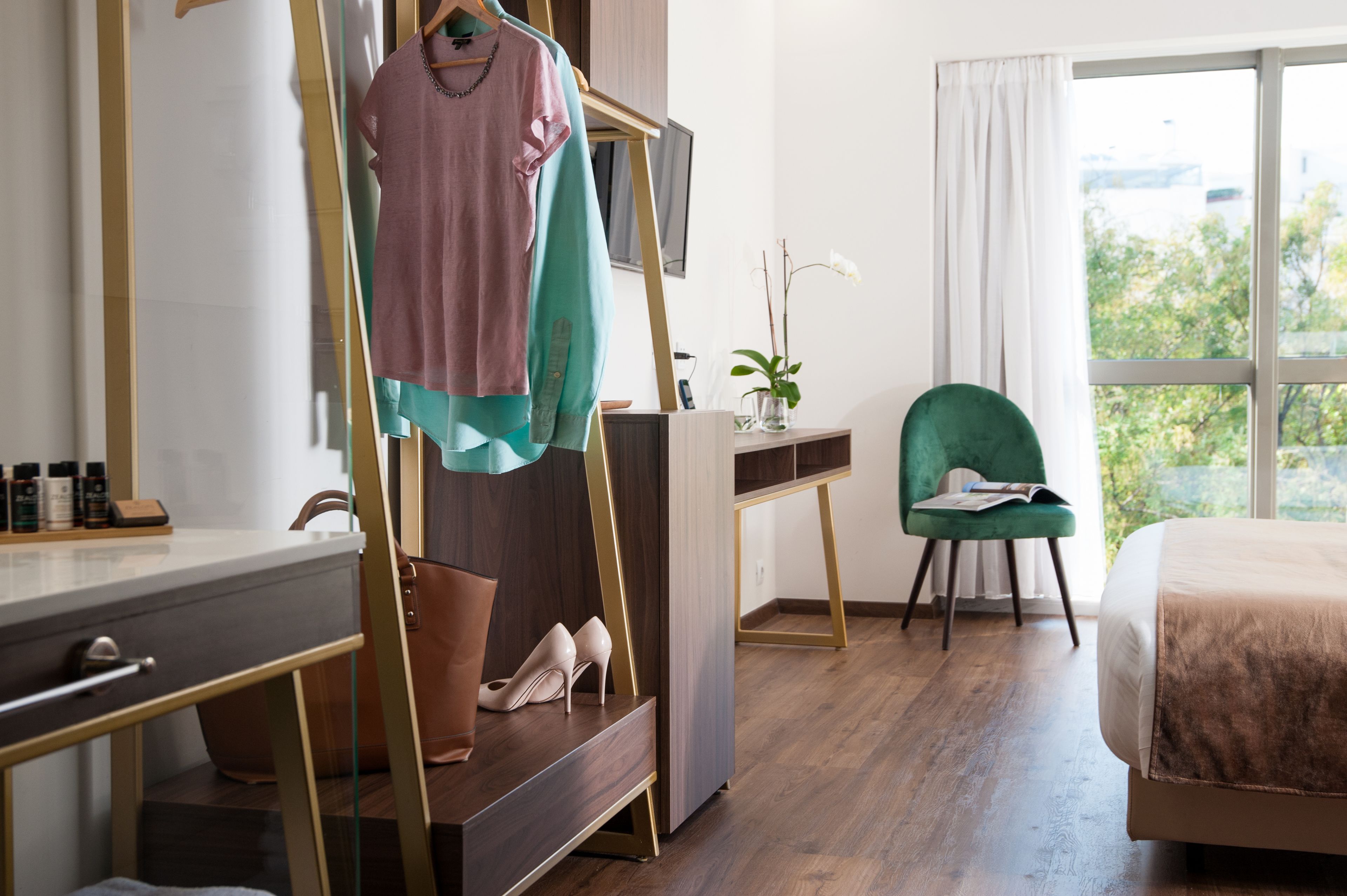 A hotel room with a bed, a desk with a chair, a hanging rack with clothes, a pair of shoes, a small table with a plant, and a window with a view of greenery.