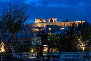 A rooftop terrace with chairs and tables, overlooking ancient ruins illuminated at dusk. - B4B Athens Signature Hotel (Athens)
