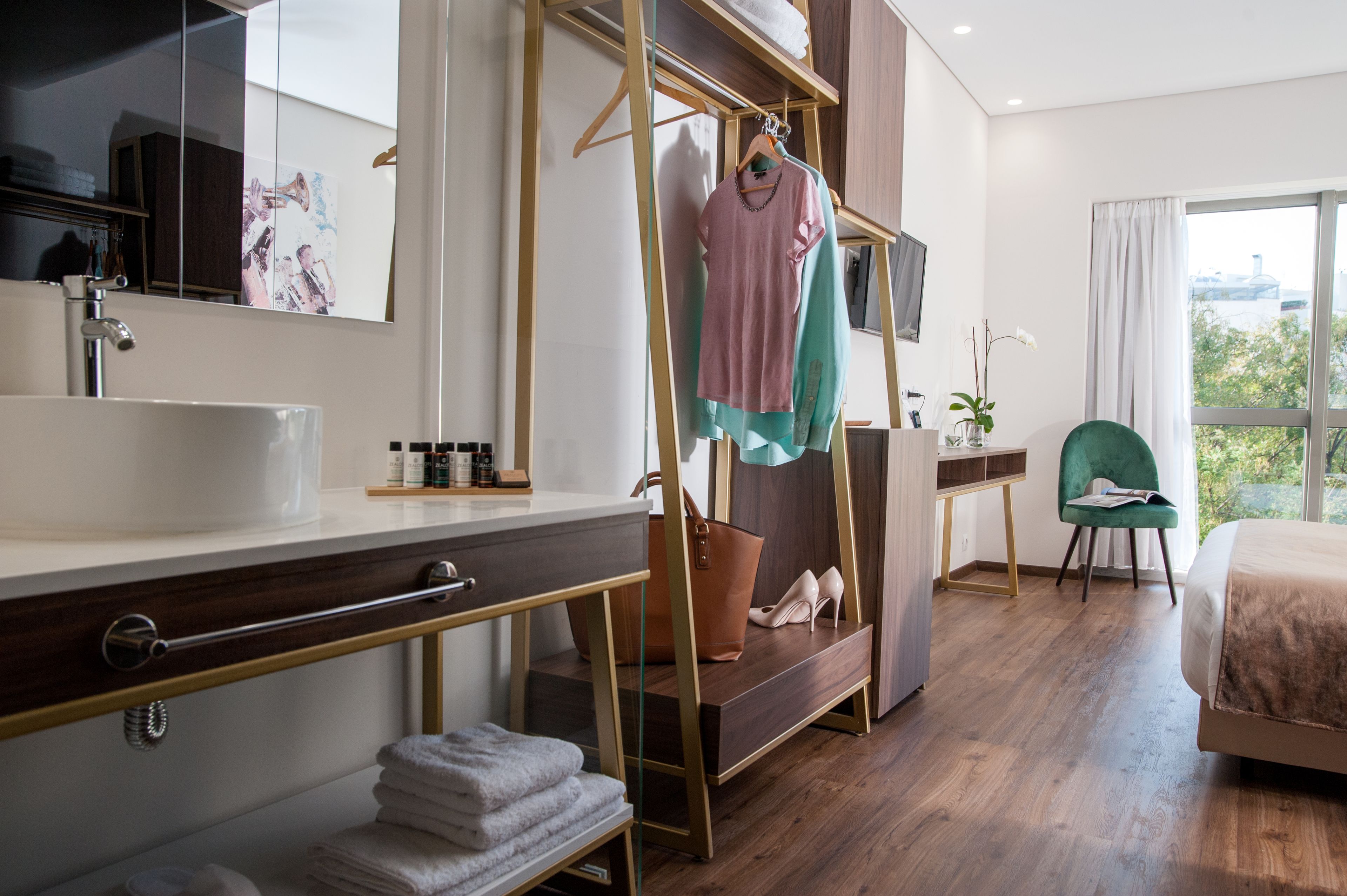 Een moderne hotelkamer met een groot raam, een houten vloer, een bed, een groene stoel, een kaptafel met spiegel en een kledingrek met kleding en schoenen.