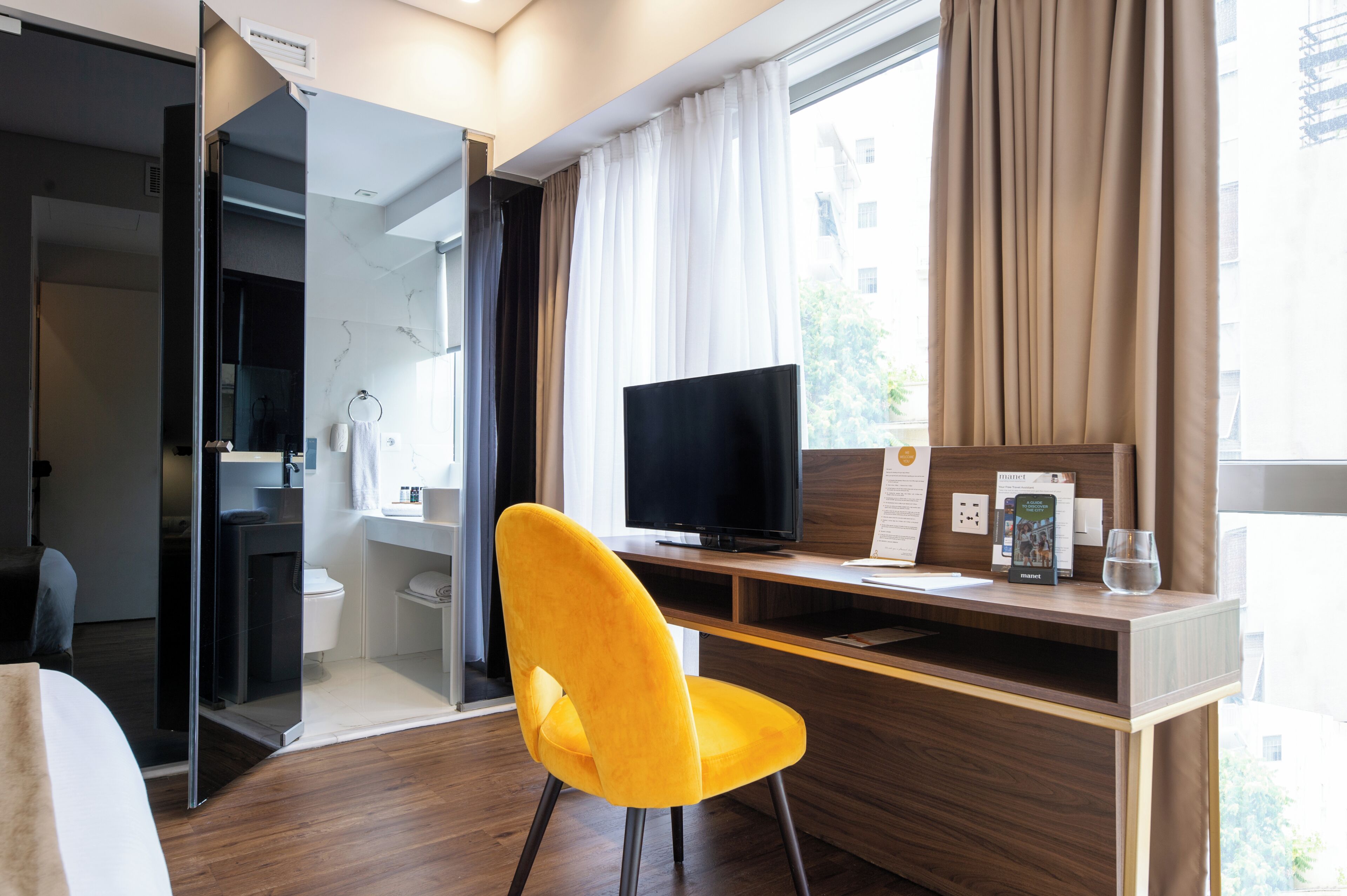 A hotel room with a wooden desk, a yellow chair, a television, a glass of water, and a window with curtains.