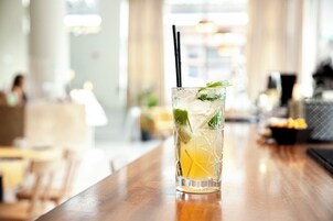 A clear glass with a straw, containing a beverage with ice and a slice of lemon, placed on a wooden bar in a well-lit indoor setting.