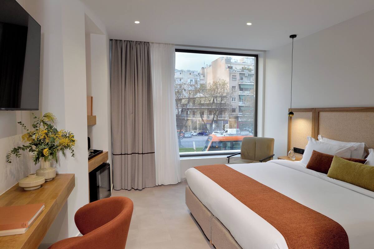 A hotel room with a large bed, a desk with a chair, a TV, a window with curtains, and a view of buildings outside.