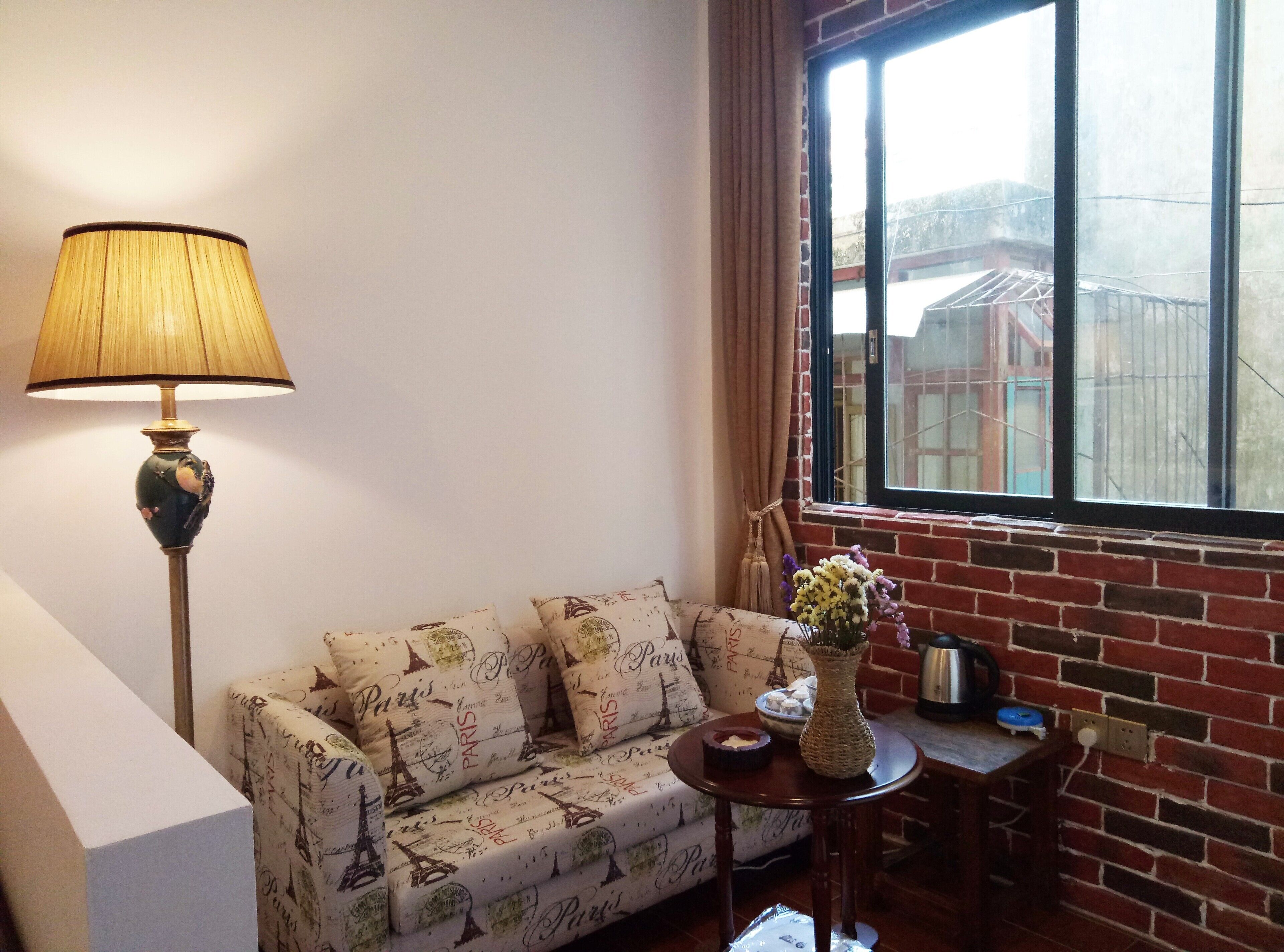 A cozy room with a sofa adorned with Paris-themed cushions, a floor lamp, a small table with a vase of flowers, and a view of a brick building through a window.