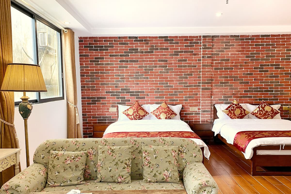 Una habitación de hotel con pared de ladrillo, dos camas con ropa de cama floral, un sofá tapizado en flores y una mesa de centro de madera con objetos decorativos.