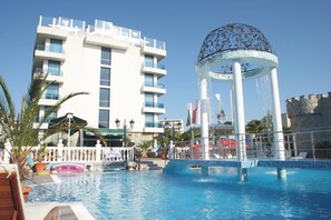 Zona de piscina de hotel con una fuente decorativa, un edificio moderno con balcones y un cielo azul despejado.