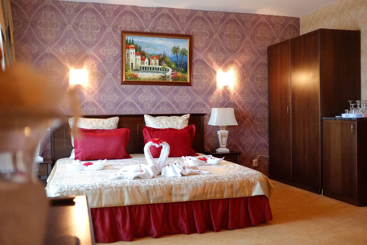 A hotel room with a bed, red pillows, a painting on the wall, and a wardrobe.