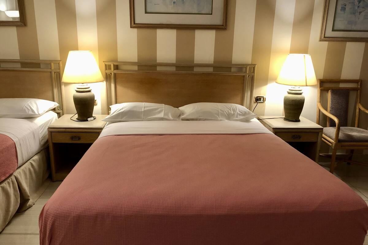 A hotel room with two single beds, a large bed, and a chair. There are lamps on the nightstands, and artwork on the walls.