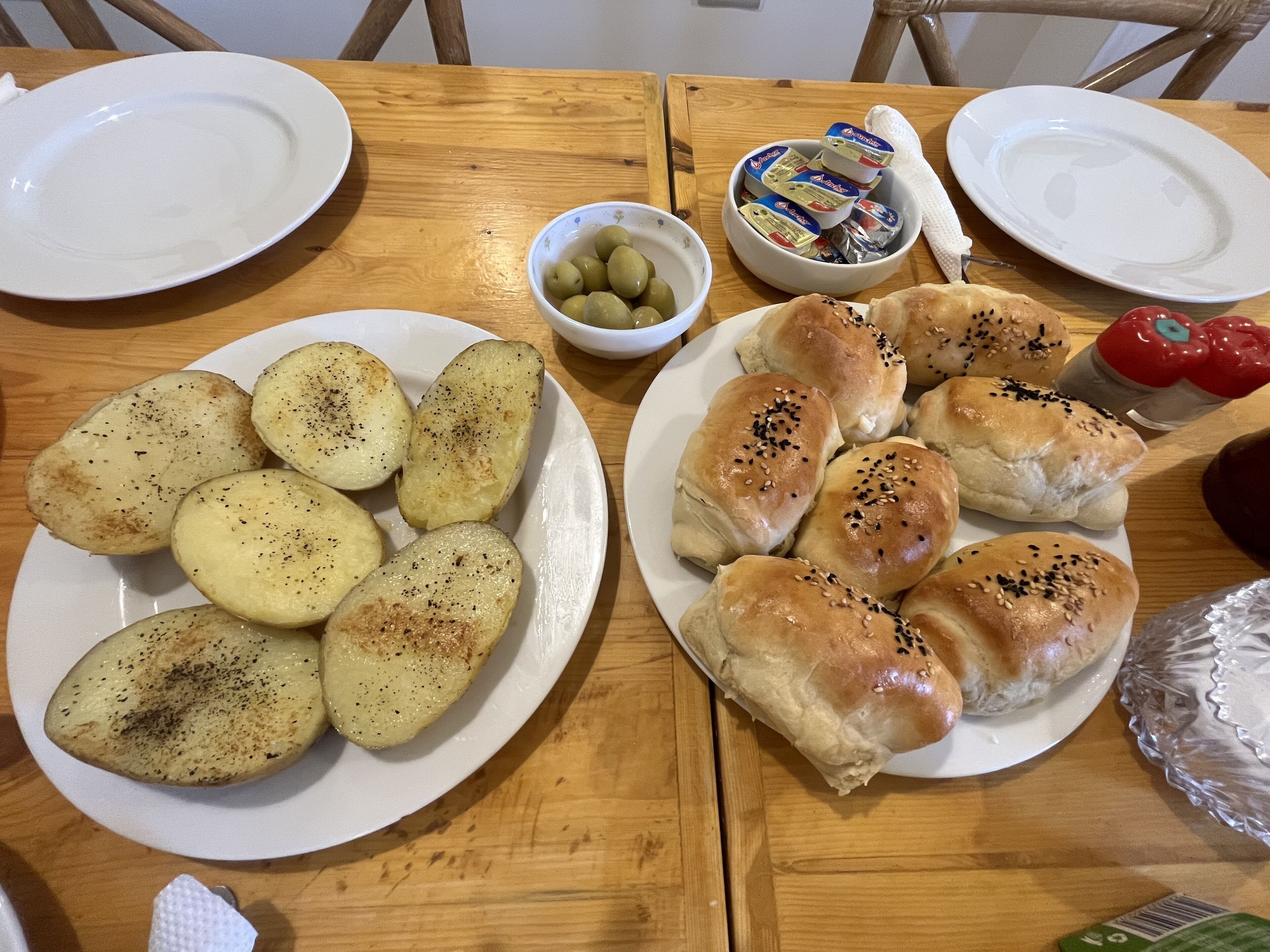 A wooden table with plates of roasted potatoes and sesame seed buns, a bowl of olives, and a container of butter.