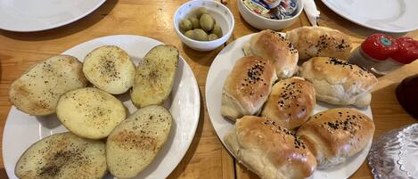 A wooden table with plates of roasted potatoes and sesame seed buns, a bowl of olives, and a container of butter.