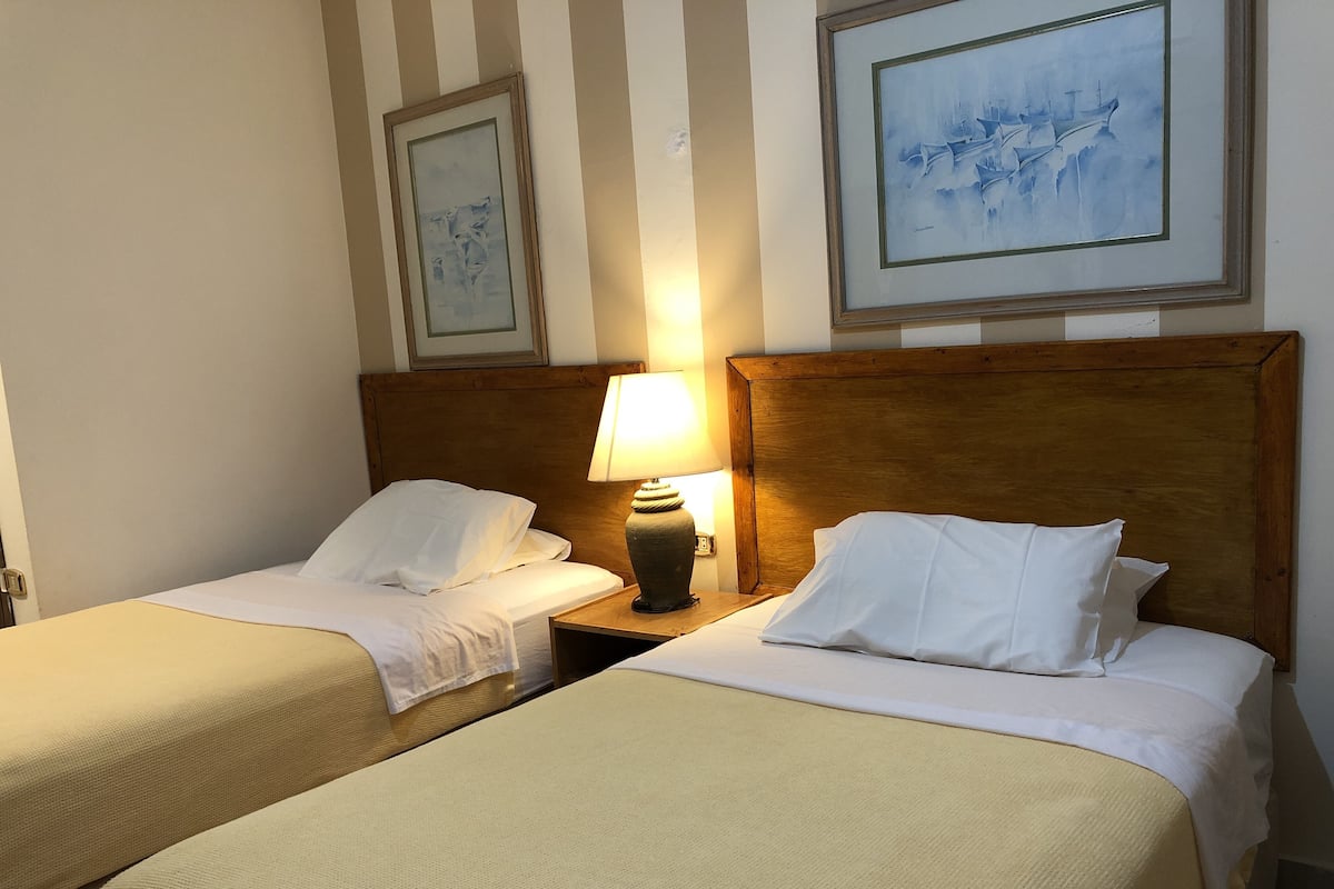 A hotel room with two single beds, a nightstand with a lamp, and two framed artworks on the wall.