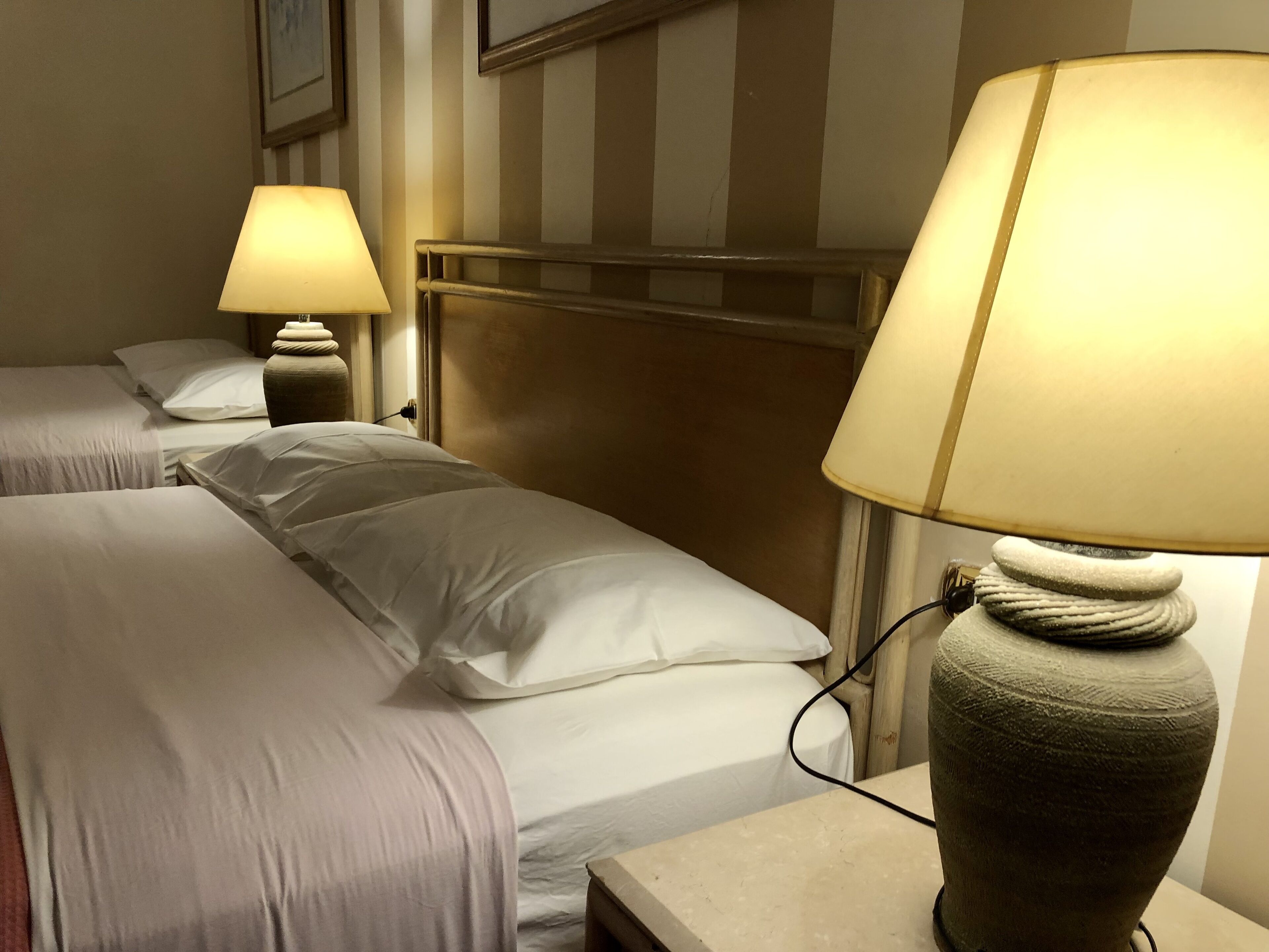 A hotel room with two beds, each with white linens and pillows, a headboard, and a bedside lamp with a textured base.