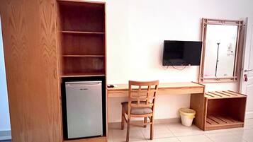 A compact room with a wooden desk, a chair, a television mounted on the wall, a small refrigerator, and a cabinet with shelves.