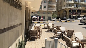 An outdoor seating area with wicker chairs and tables, a tiled floor, and a view of the street with parked cars and buildings in the background. - Masaya Suites (Hurghada)