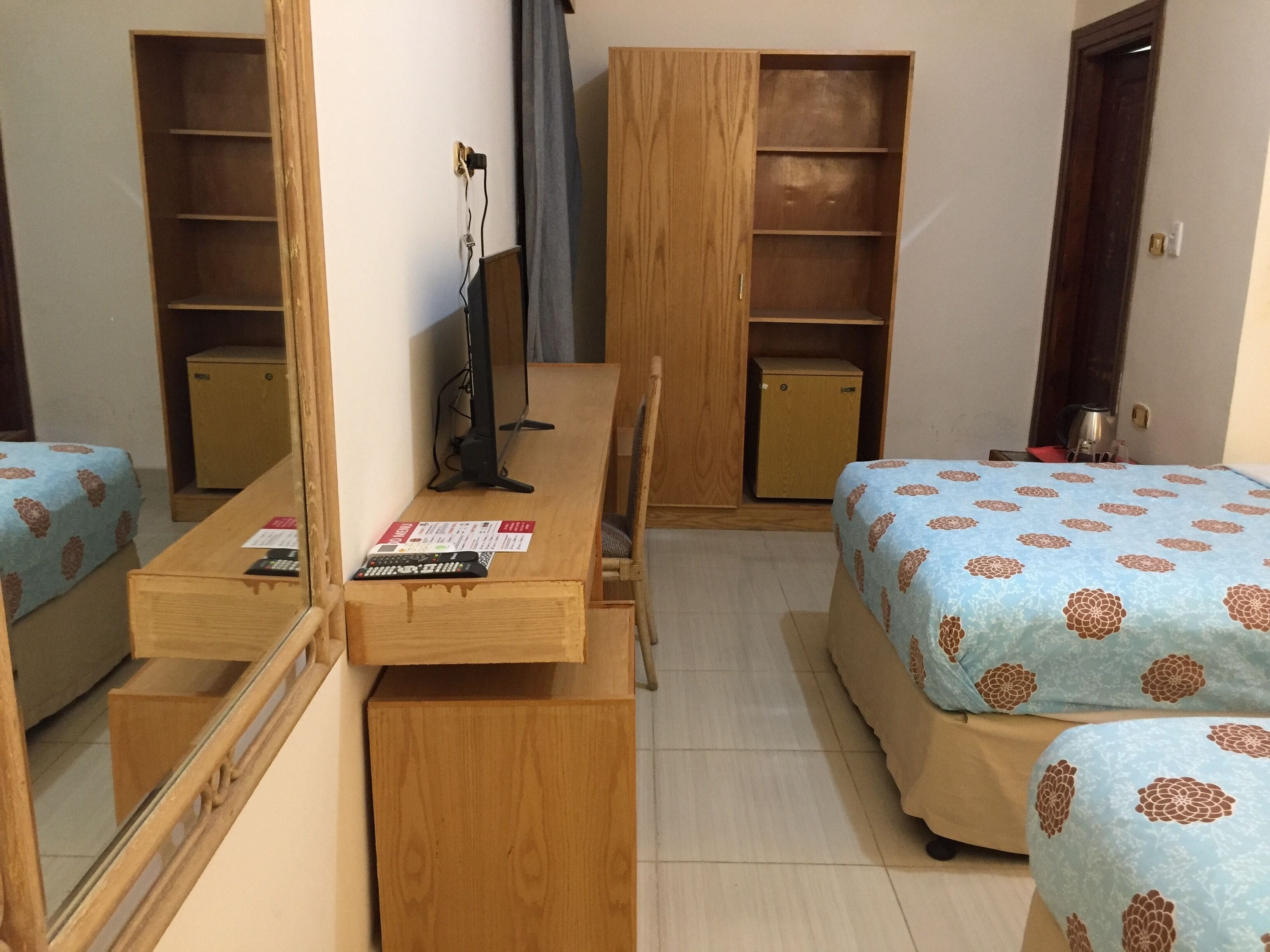 Habitación de hotel compacta con dos camas individuales, un escritorio de madera con silla, un armario pequeño y un espejo.