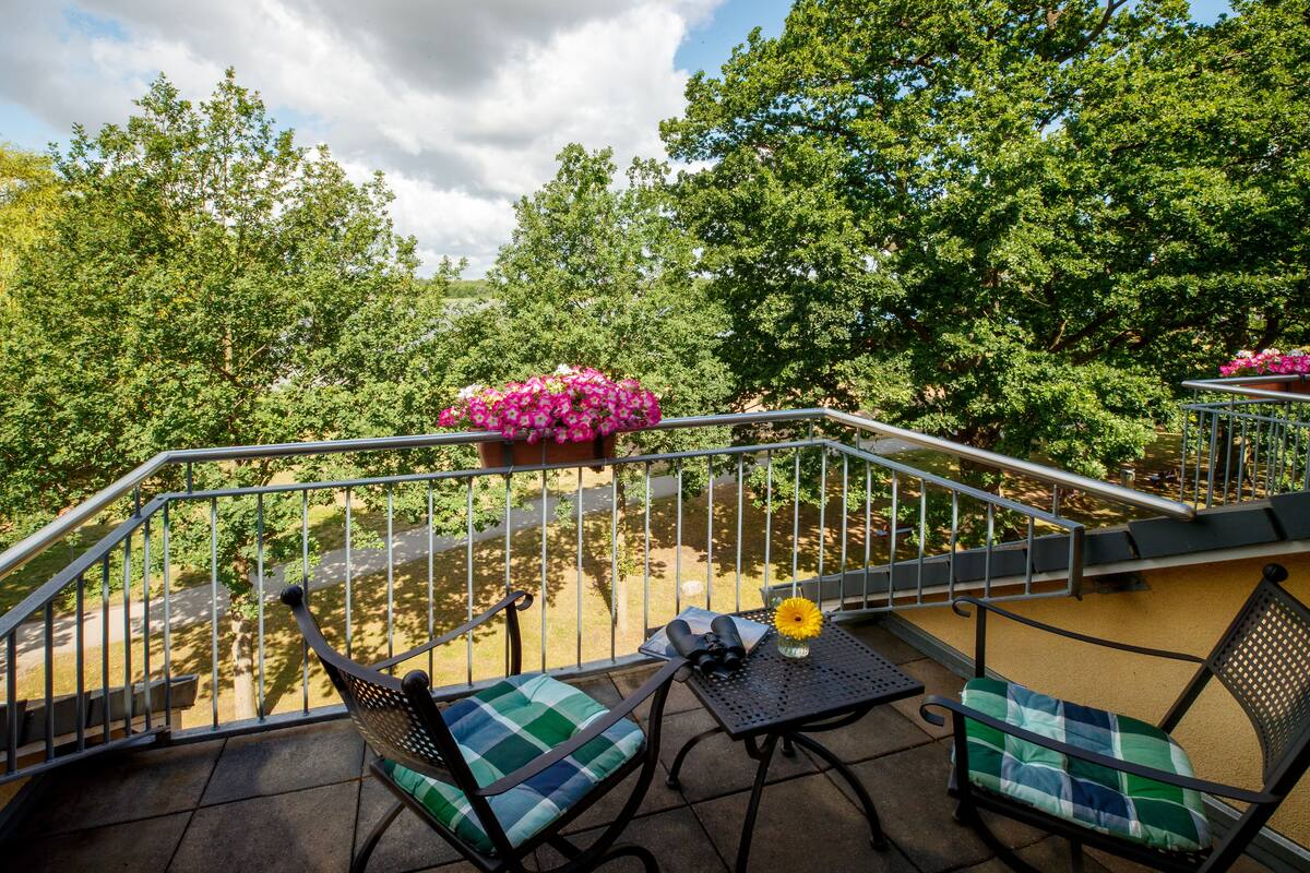 A balcony with two chairs, a table, and a railing overlooking a garden with trees and a pond.