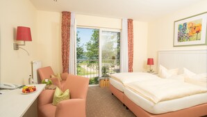 A hotel room with a bed, a desk with a phone, a chair, a small table with fruit, and a view of the outdoors.