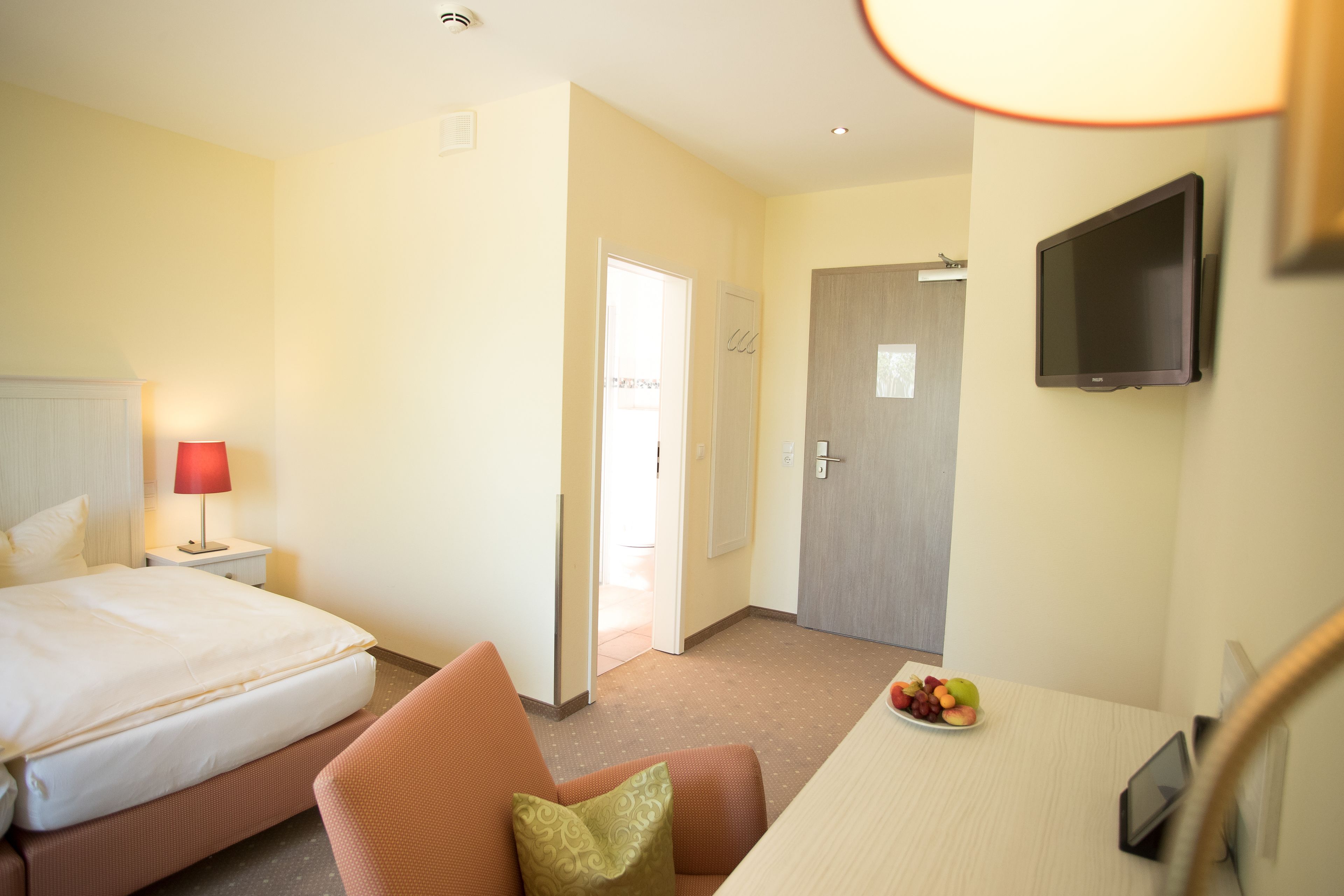 A hotel room with a bed, a desk with a fruit bowl, a TV, and a door leading to another room.