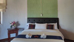 Une chambre à coucher avec un toit de chaume, des murs en briques vertes, un lit avec une literie blanche et des oreillers à carreaux, une table de chevet avec un vase de fleurs et des lampes fixées au mur.