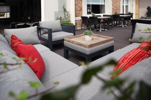 Un área de estar al aire libre con sofás grises y cojines rojos, una mesa de centro de madera con una planta, y un muro de ladrillo al fondo.