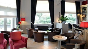 Una sala de estar moderna con un sofá, sillones y una mesa de centro. Hay dos ventanas con cortinas, una lámpara y una estantería.