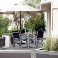 Un patio al aire libre con una mesa y sillas bajo un paraguas grande, rodeado de follaje y un muro de ladrillo.
