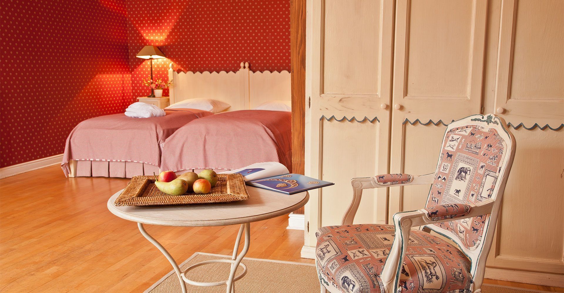 A bedroom with two beds, a small table with fruits and a book, and a patterned chair.