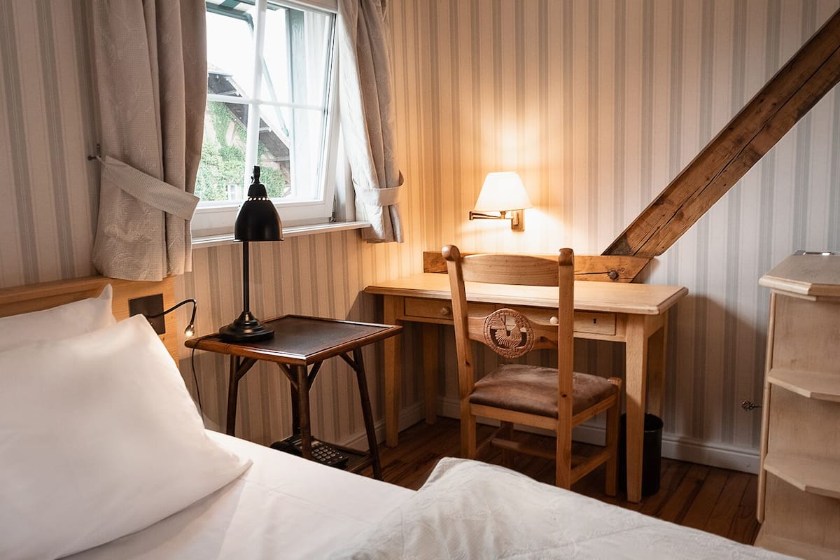 A room with a bed, a desk with a lamp, a chair, and a window with curtains.