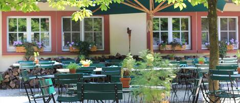 An outdoor seating area with green metal chairs and tables, surrounded by potted plants and a building with large windows.