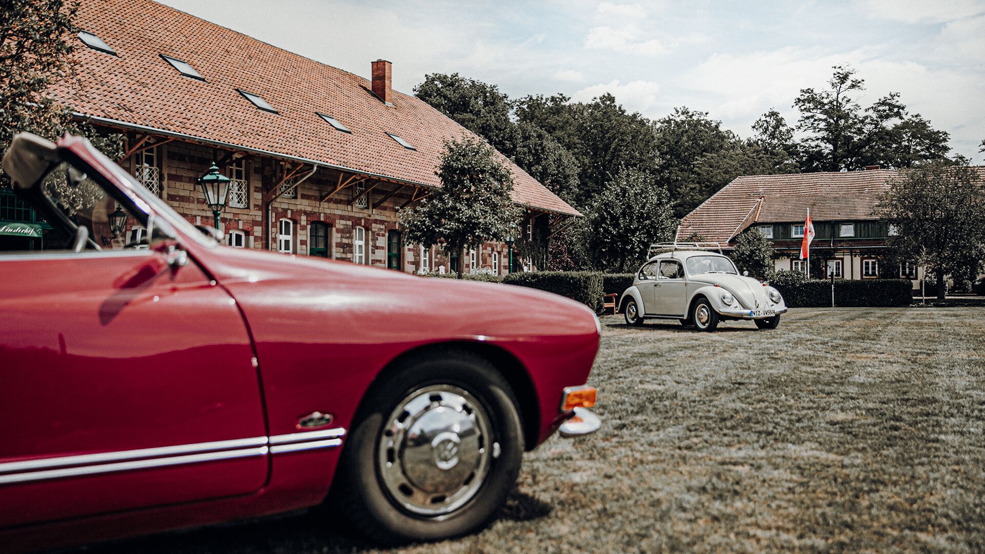 A red vintage car parked in front of a building with a tiled roof and a white vintage car in the background.