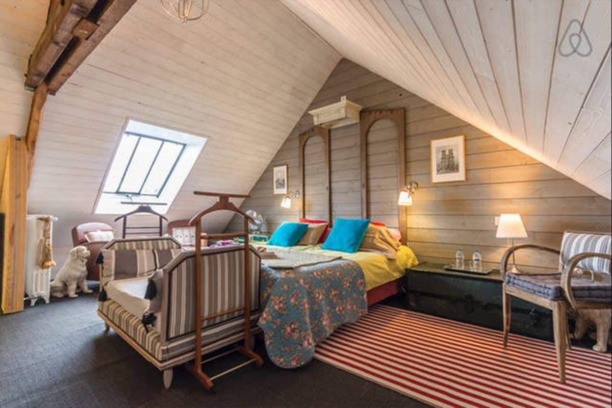 A cozy attic bedroom with a sloped ceiling, a bed with colorful bedding, a wooden chair, a small table, and a dog.